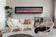 Load image into Gallery viewer, Napoleon Entice linear electric fireplace wall-mounted above a sofa in a bright living room with a hanging chair