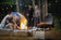 Load image into Gallery viewer, Friends gathered around a campfire with the Napoleon TravelQ 285 blue portable grill cooking chicken nearby