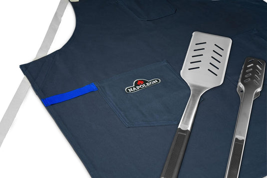 Navy Napoleon anniversary apron displayed with a grilling spatula and tongs