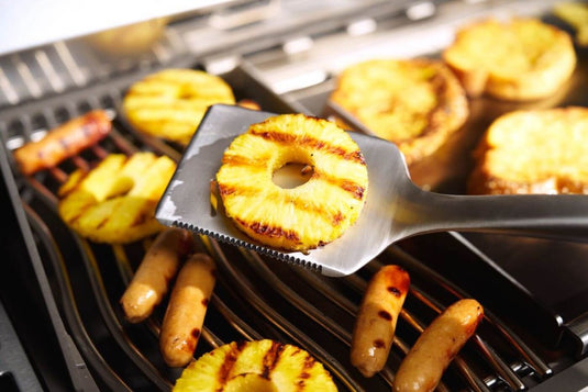 Napoleon GATL007 stainless steel grill spatula lifting a grilled pineapple slice over a barbecue grill