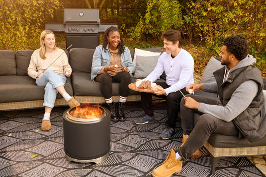 Four people enjoying an outdoor gathering around the Napoleon Timberwolf 18-inch matte black smokeless fire pit