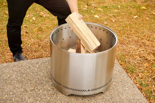 Person placing firewood into the Napoleon Timberwolf 18-inch stainless steel smokeless fire pit (NWFPMTI-SS) outdoors.